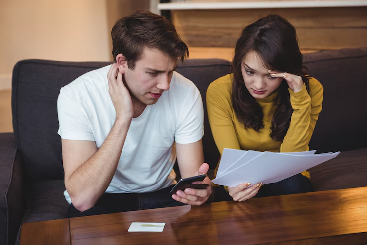 A worried couple sits on a couch reviewing financial documents, with the man holding a smartphone and the woman holding papers — both displaying expressions of concern or stress, possibly over bills or debt issues.