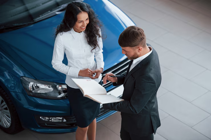 Salesperson and customer standing beside a blue car in a showroom, reviewing documents together with the customer smiling, indicating a possible car purchase or lease agreement."