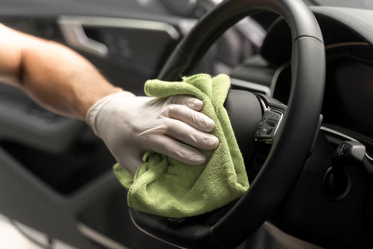 "Person wearing a glove cleaning a car steering wheel with a green microfiber cloth, focusing on disinfecting or detailing the vehicle interior."
