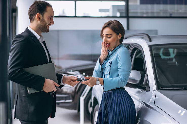 A smiling car salesman hands over car keys to a surprised and excited woman standing next to a new car inside a showroom, symbolizing a joyful car purchase experience.