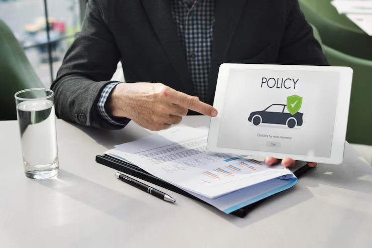 "Businessperson pointing at a tablet screen displaying a car insurance policy with a green shield checkmark icon, alongside documents, a pen, and a glass of water on the table."