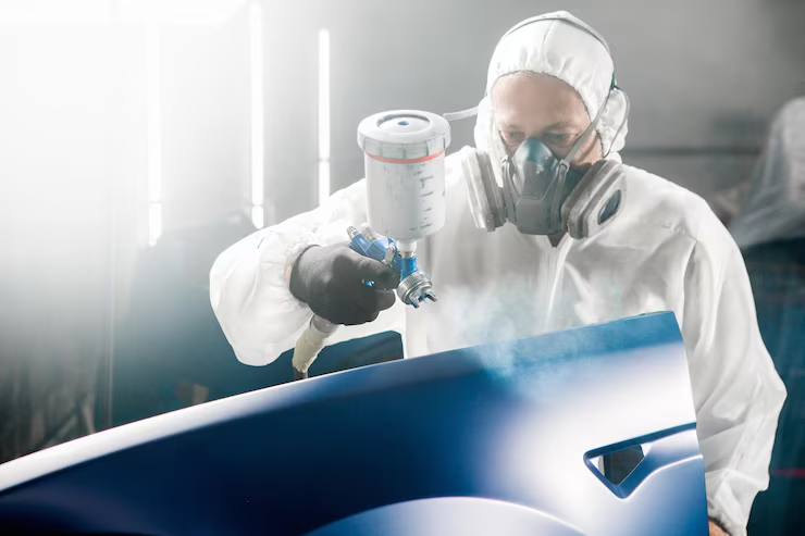 "Auto body technician wearing protective gear and a respirator while spray painting a blue car door in a professional workshop."