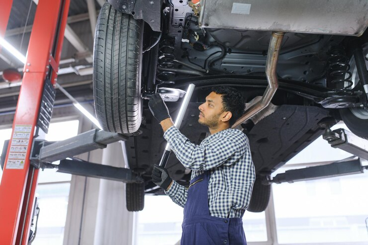 "Auto mechanic inspecting the underside of a raised vehicle in a garage, using a flashlight and wearing a checkered shirt and overalls."