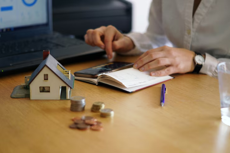 A person calculates finances at a desk with a notebook, coins, a model house, and a laptop, representing budgeting, mortgage planning, or financial management related to homeownership.