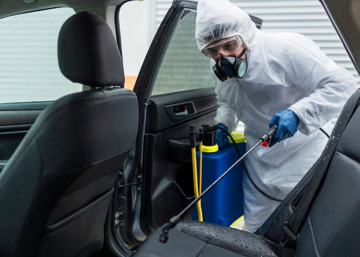 "A person in full protective gear and respirator disinfecting the interior of a car using a spray nozzle attached to a blue sanitation tank."