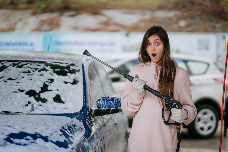"Surprised woman in a pink hoodie holding a pressure washer while cleaning a foam-covered blue car at a car wash."