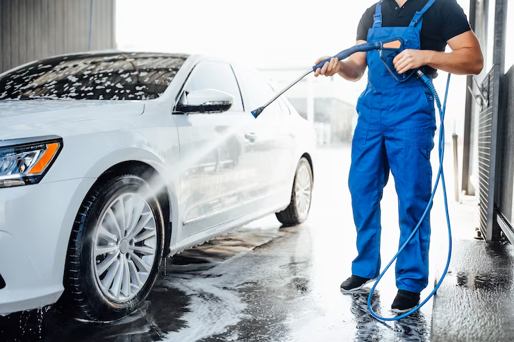 Man in blue overalls pressure washing a white car at an auto wash facility.