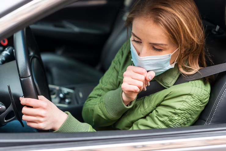 A woman wearing a green jacket and a surgical mask is sitting in the driver's seat of a car, coughing into her hand, possibly indicating poor air quality or illness.