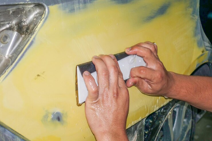 Close-up of a person's hands sanding a yellow car surface with a sanding block, preparing the area for painting or refinishing during the auto body repair process.