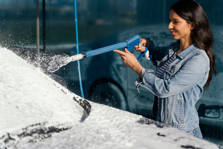 A woman wearing a denim jacket sprays foam on a car windshield using a pressure washer at a car wash station, smiling as she cleans the vehicle.