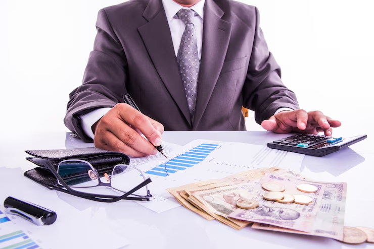 A businessman in a suit analyzing financial charts with a pen, using a calculator on a desk covered with Indian currency notes, coins, documents, glasses, and a wallet.