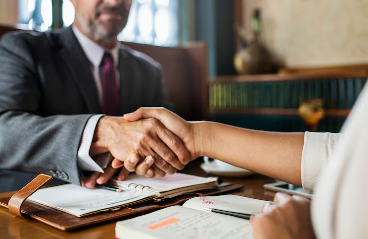 Two professionals shaking hands across a table with open notebooks and documents, indicating a business agreement or contract discussion.