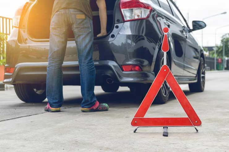 A red emergency warning triangle is placed on the ground behind a car with its trunk open, while a person stands next to the vehicle, possibly dealing with a roadside issue or breakdown.