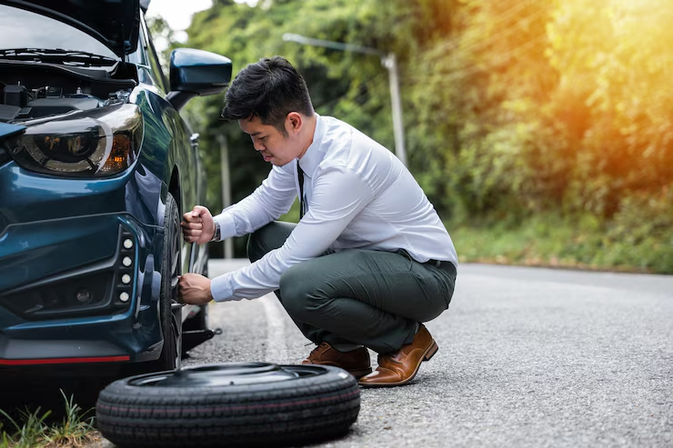 Man in formal attire kneeling on the roadside while changing a flat tire on a dark blue car, with the hood open and spare tire placed nearby.
