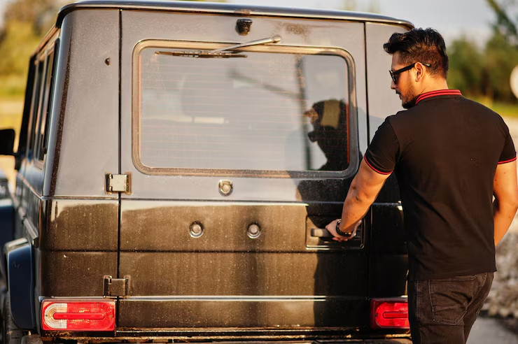 Man in a black shirt and sunglasses opening the dusty rear door of a black SUV during sunset.