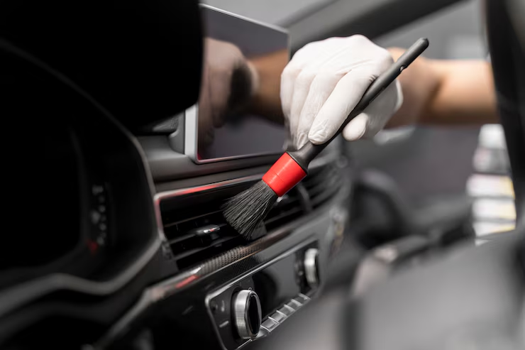 Person wearing a glove cleaning a car’s air vent using a detailing brush inside a vehicle interior.