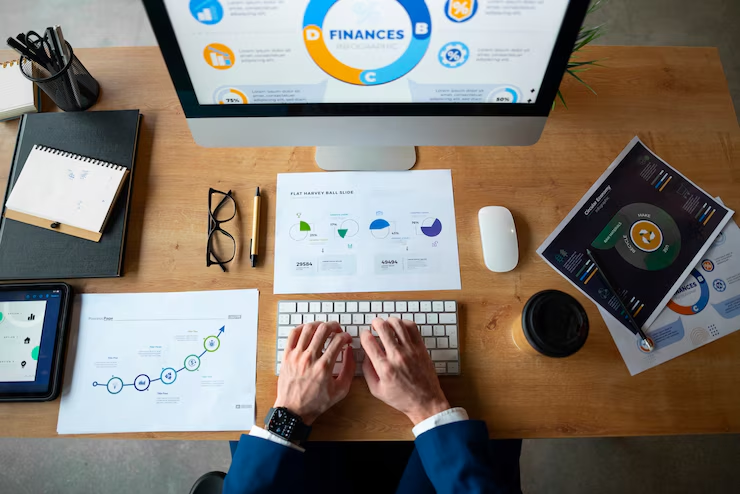 Overhead view of a businessperson typing on a keyboard at a wooden desk with a computer displaying financial data, surrounded by charts, graphs, a notebook, coffee cup, glasses, and tablet.