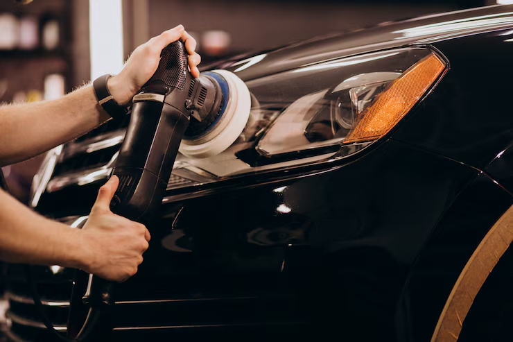 Person polishing the front headlight area of a black car using a power buffer tool in a detailing or auto workshop setting.