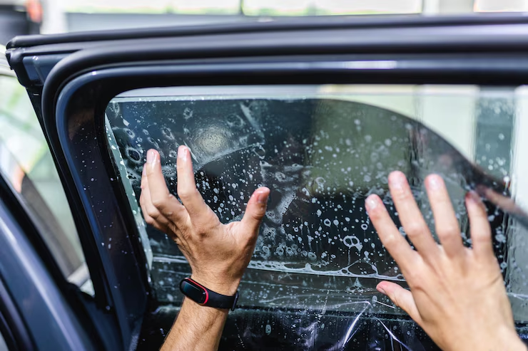 Person applying a window tint film to a car window using both hands, with soap and water solution visible on the glass surface.