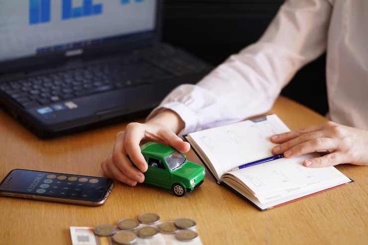 Person sitting at a desk holding a small green toy car, with a notebook open, coins on the table, a smartphone displaying a calculator app, and a laptop in the background.