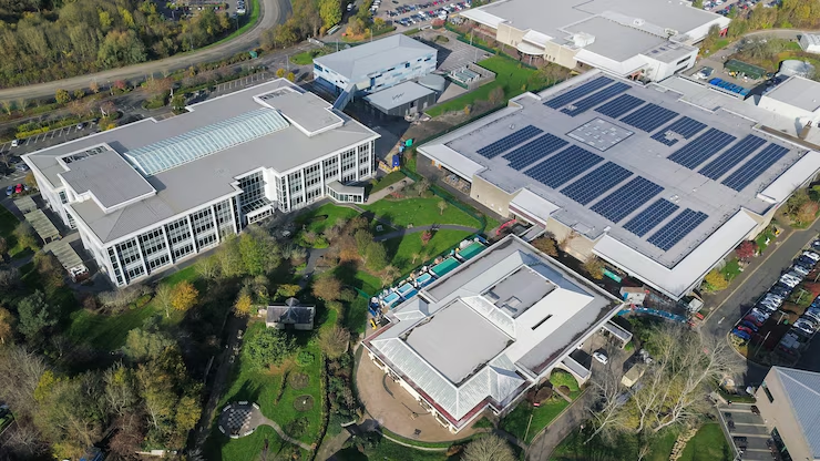 Aerial view of a modern campus or business complex featuring multiple buildings, solar panels on rooftops, landscaped gardens, and parking areas surrounded by trees.