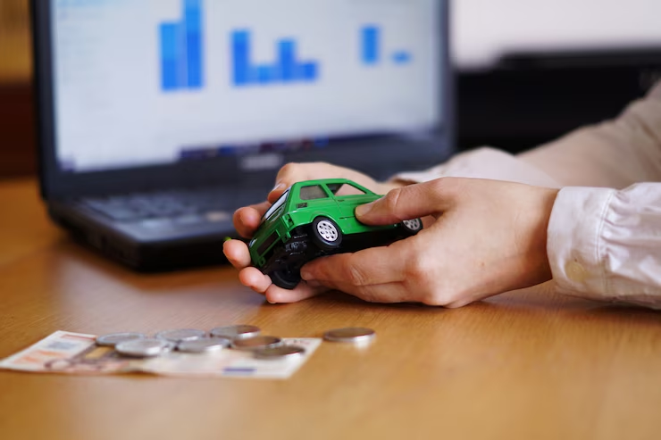 Close-up of hands holding a green toy car with coins and Euro banknotes on a wooden table, and a laptop displaying bar charts in the background, representing car finance or insurance concepts.