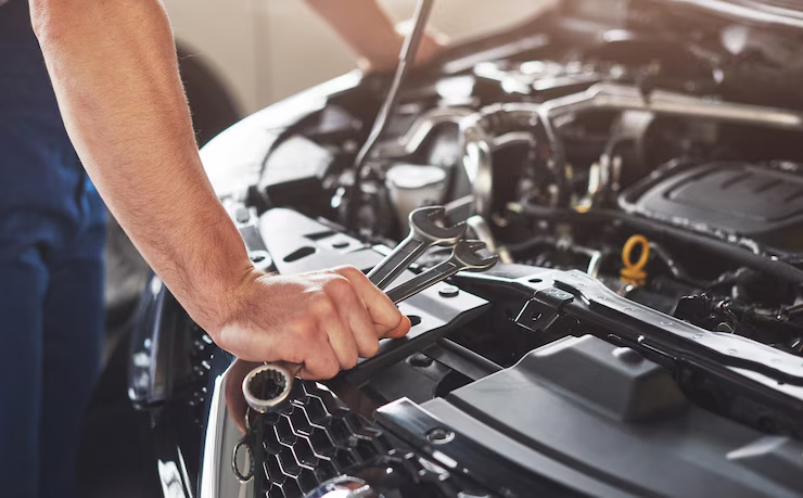 Mechanic holding wrenches while working under the hood of a car, showcasing vehicle maintenance or repair in a garage setting.