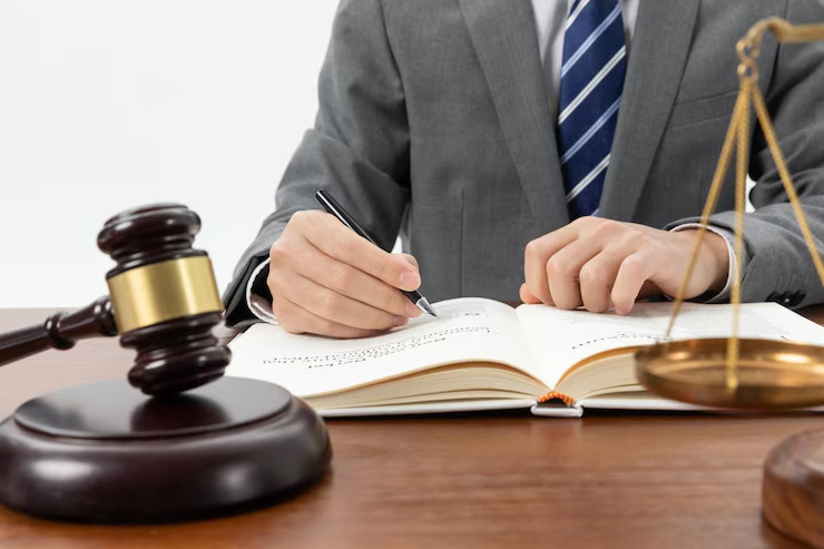 A person in a suit writing in a book at a desk with a judge's gavel and justice scale, symbolizing legal work, court proceedings, or law practice.