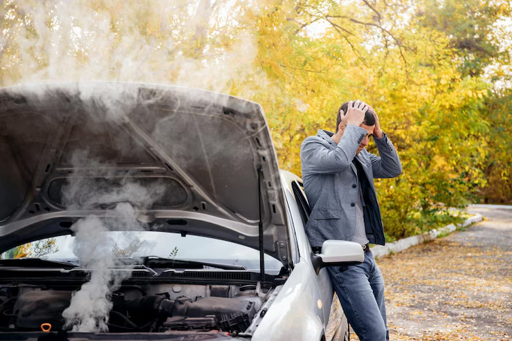 Man standing next to a car with the hood open and smoke coming out of the engine, holding his head in frustration, indicating a vehicle breakdown or engine overheating problem.