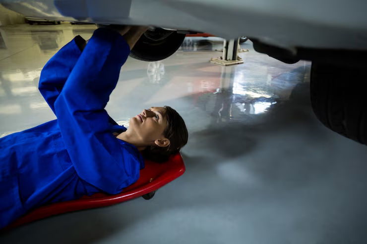 A female mechanic in a blue uniform lies on a red creeper while working underneath a car in a clean, well-lit auto repair shop.