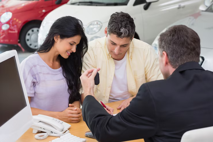 A couple signing paperwork at a car dealership desk while a salesperson hands over a car key, indicating the final steps of purchasing or leasing a vehicle.