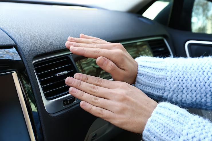 Hands in a light blue sweater held near a car's air vent, checking or warming up with air coming from the dashboard ventilation system.
