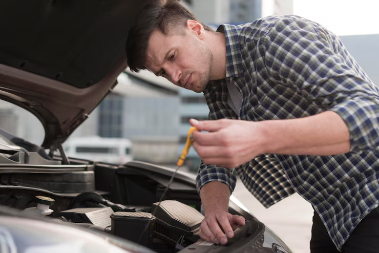 A man in a plaid shirt checking the engine oil level using a dipstick under the hood of a car, appearing focused while performing vehicle maintenance.