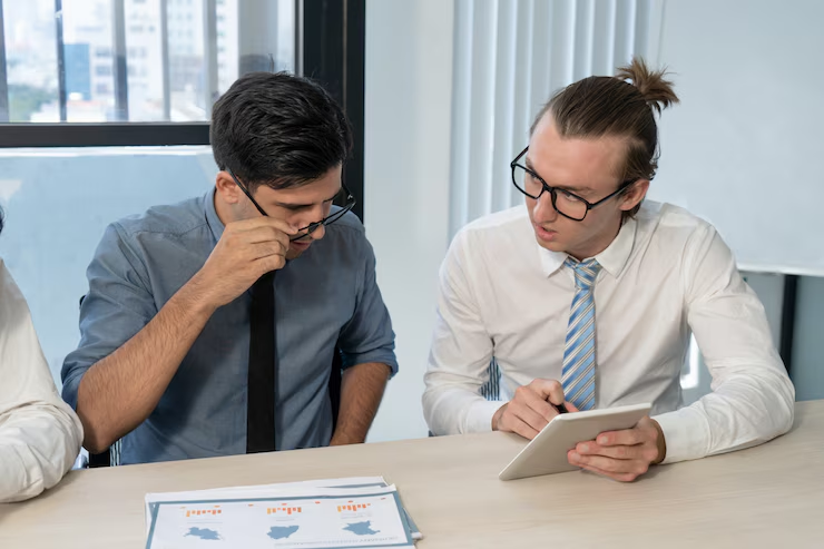 Two men in business attire are seated at a desk in an office, discussing data displayed on a tablet. One man is holding the tablet while the other adjusts his glasses to look at printed charts and graphs on the table.