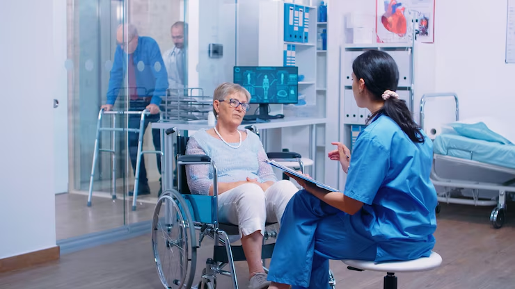 A nurse in blue scrubs sits and consults with an elderly woman in a wheelchair inside a medical facility. In the background, two elderly men with walkers and a doctor are visible through a glass partition, indicating a clinical setting focused on senior care.
