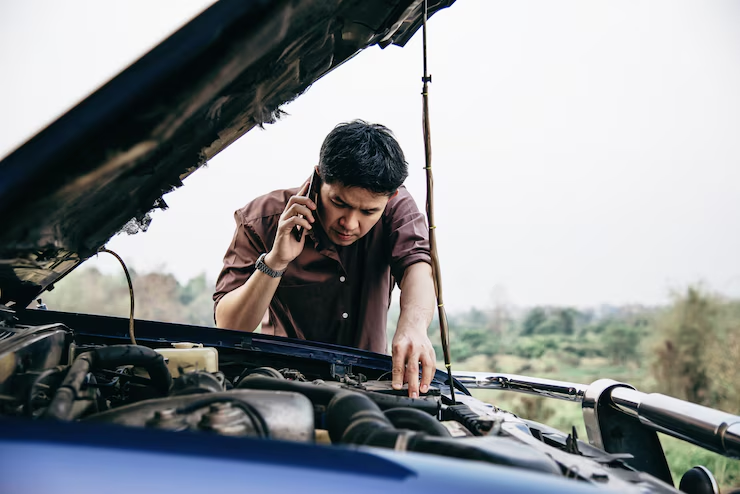 A man is inspecting the engine of his car with the hood open while talking on the phone, likely calling for roadside assistance due to a car breakdown. The scene is set in a rural or remote area with greenery in the background.