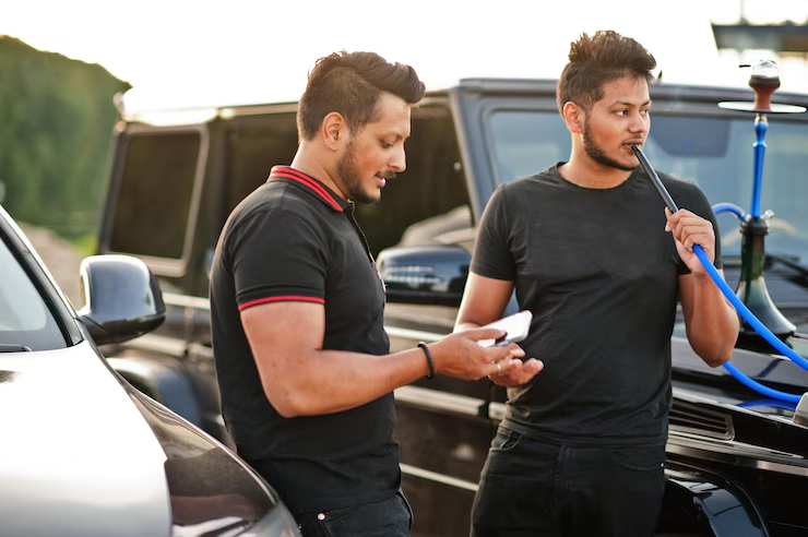 : Two men standing next to a black car. One is holding a phone and the other is smoking from a hookah, both dressed in casual black shirts, with a relaxed setting outdoors.