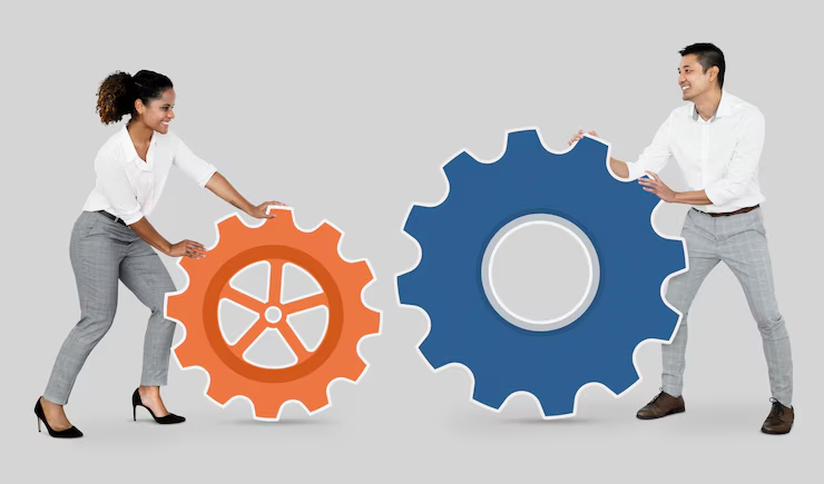 The image shows two individuals holding large, symbolic gears, one orange and the other blue. The man and woman are interacting with these oversized gears, which symbolize teamwork, collaboration, or operational efficiency. The image uses the gears to visually represent the concept of mechanisms working together, highlighting the importance of coordination and collaboration. The background is plain, making the focus clearly on the people and the gears.