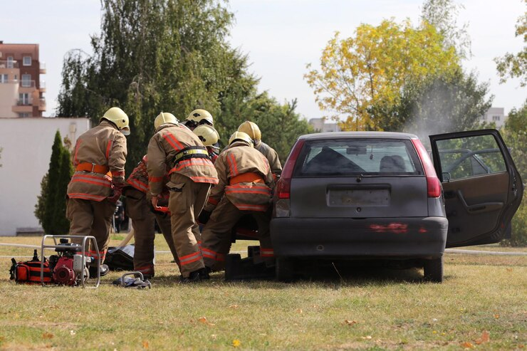 The image shows a team of firefighters in full gear responding to an emergency. They are working together to assist someone involved in a car accident. The firefighters are likely helping to remove the person from the vehicle or performing other emergency rescue operations. The background suggests the scene is taking place outdoors, with trees and buildings visible. The car has an open door, and some smoke is visible, possibly indicating the aftermath of a crash or a vehicle fire. The teamwork and urgency are evident in the firefighters' coordinated efforts.
