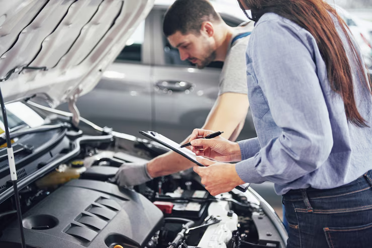 "Auto mechanic working under the hood of a car while a woman with a clipboard takes notes during a vehicle inspection or service evaluation."