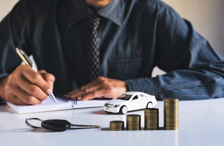 "Businessman in formal attire writing in a notebook with a toy car, car key, and stacked coins arranged in ascending order on the table, symbolizing car finance or auto loan planning."