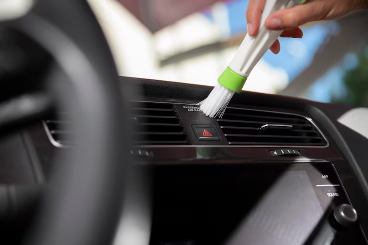 "Close-up of a person using a small brush to clean the air vent of a car dashboard, focusing on interior detailing and maintenance."