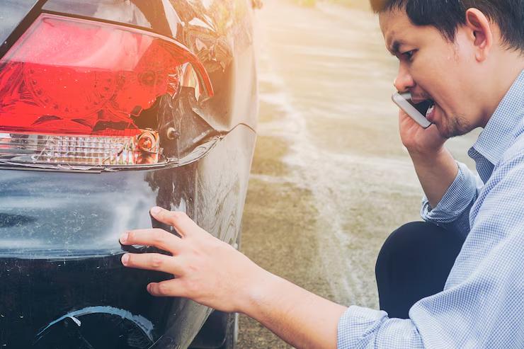"Man inspecting damage to the rear bumper and taillight of his car while talking on the phone, likely reporting an accident or contacting insurance."