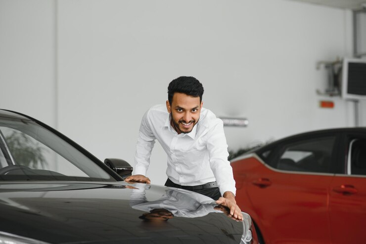 "Smiling man in a white shirt inspecting or admiring a black car in a showroom, with another red car in the background, possibly indicating a car buying experience."