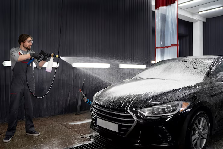 "Man using a high-pressure foam sprayer to wash a black car at an indoor car wash facility, with soap suds covering the vehicle’s hood and windshield."