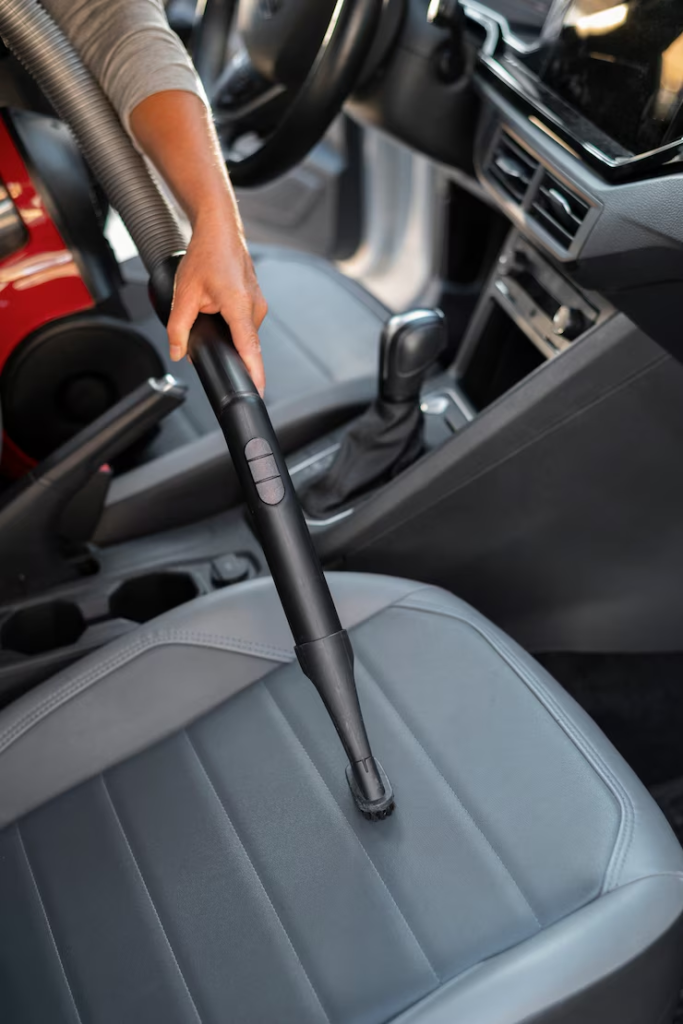 "Close-up of a person vacuuming the front seat of a car interior using a narrow brush attachment, focusing on detailed vehicle cleaning."