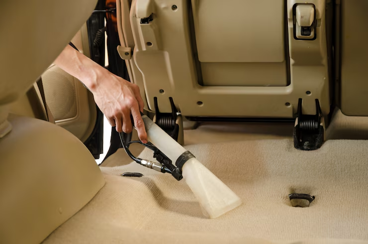 "Person using a professional extractor vacuum to clean the beige carpet of a car interior with the rear seats folded forward."