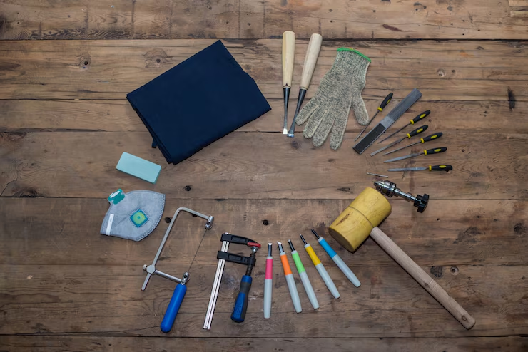 "Various hand tools arranged on a wooden surface, including files, chisels, hammers, screwdrivers, gloves, a saw, face mask, apron, and carving tools, suggesting a setup for woodworking or craftwork."